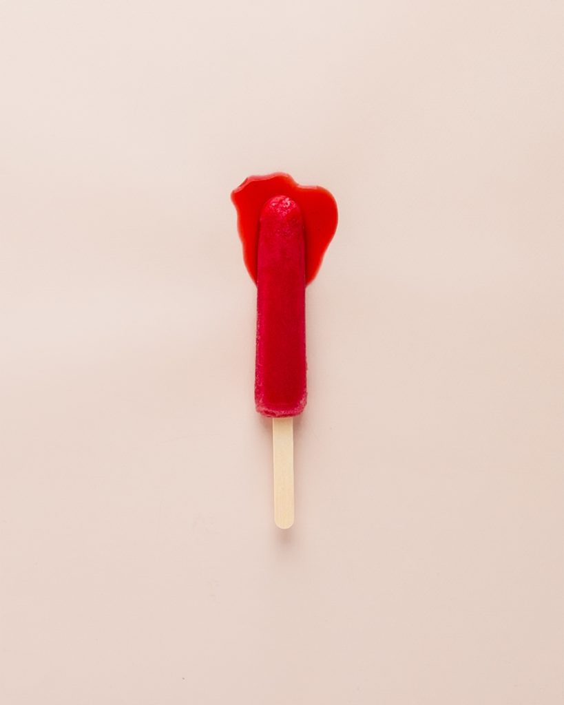 Schmelzendes Eis am Stil vor einem rosa Hintergrund, es soll den Tampon, einen konventionellen Monatshygieneartikel, darstellen
