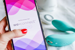 Die We-Vibe Sync App we-connect fuer Sex in Fernbeziehung ist auf dem Handy geoeffnet. Im Hintergrund der We-Vibe Sync in tuerkis